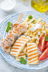 Slices of grilled haloumi cheese, souvlaki skewers, greek-style fries and tomatoes served on a blue and white plate, vertical shot, close-up