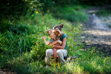 Mom and son in park among grass are playing fishing with stick and fishing rod. Mom hugs boy, helps...