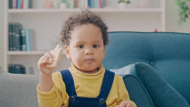 Funny curly-haired toddler eating snack while playing at home, happy childhood