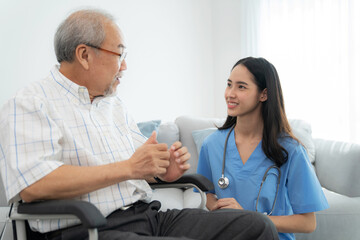 Attractive caregiver nurse sitting beside elderly man who sitting on the wheelchair and smile at home. Doctor take care and encourage  patient. Rehabilitation and healthcare concept.