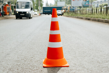 Close-up of road safety cone on asphalt, cars in bokeh. Striped orange and white warning sign