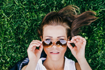 Close up portrait of beautiful young woman laying on green grass and looking over her aviator...