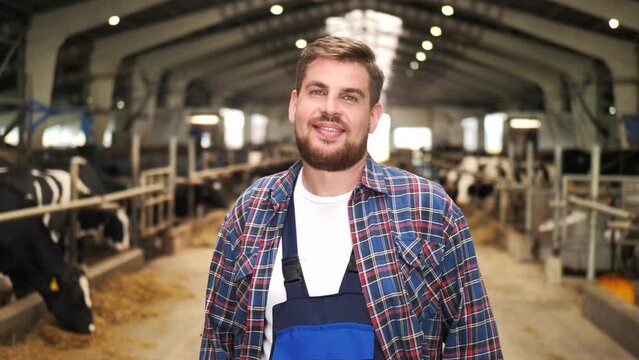 Young smiling farmer in cowshed looks around and gives thumbs up