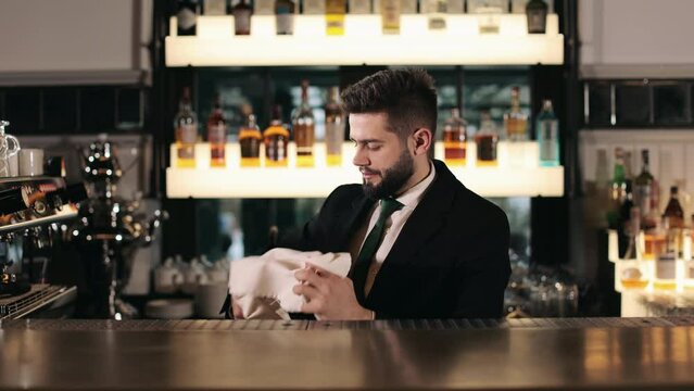 Handsome young bartender wiping wine glass with towel behind bar counter. Caucasian man in black suit preparing silverware for serving at modern restaurant.