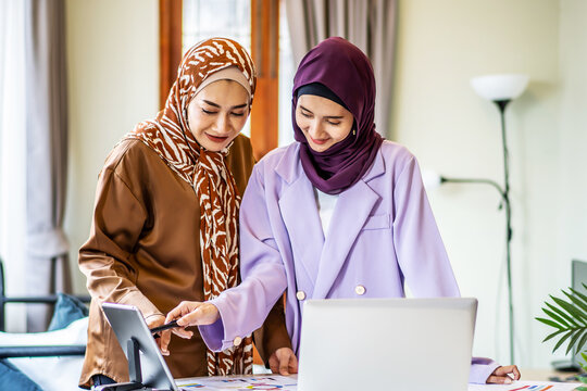 Two Asian muslim women wearing hijab working together with using laptop pc tablet and paper showing graphic chart in living room at home, respect and collaboration concept.