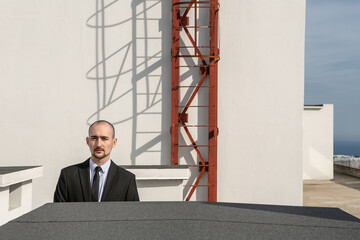 a man in a black suit with a tie stands against the backdrop of a fire escape on the roof of a house