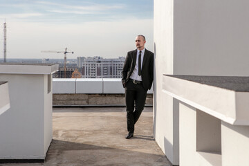 a man in a black suit with a tie leaned against the wall on the roof against the background of the panorama of the city and building cranes