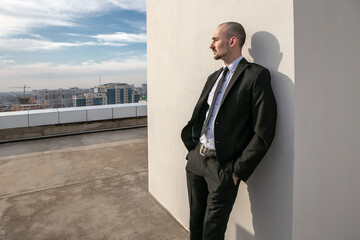 a man in a black suit with a tie leans against the wall and looks at the panorama of the city