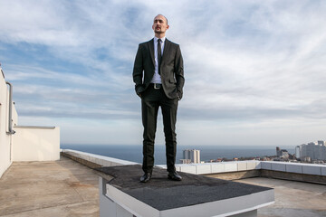 a man in a black suit and tie stands on a roof with the sea skyline in the background