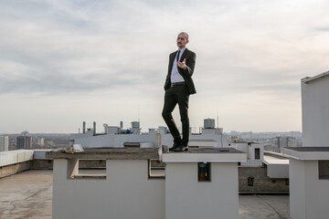 a man in a black suit with a tie stands on the roof and holds out his hand in front of him