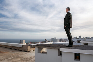 a man in a black suit with a tie stands on the roof and looks towards the horizon at the sea