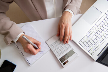 businesswoman counts the budget and makes notes in a notebook on the desktop with a calculator and a laptop.