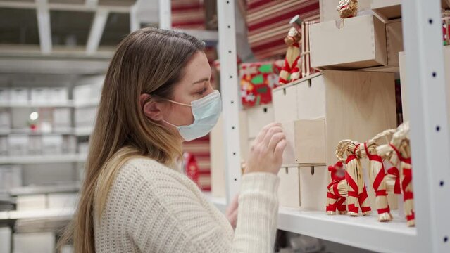 a woman in a protective mask looks at a wooden box with retractable shelves, next to it on the shelf is a Christmas goat made of straw, decorated with a red ribbon