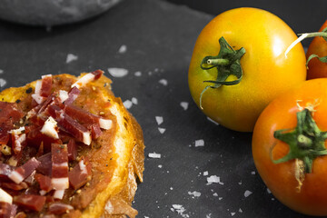 Ham toast with tomato, oil and pepper on a slate plate placed next to rustic wooden kitchen utensils, salt shaker and recipe ingredients such as tomato and broad beans