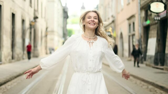 Attractive middle aged woman in white dress walking down the street and looking ahead. Happy relaxed lady walking on the city centre enjoying.