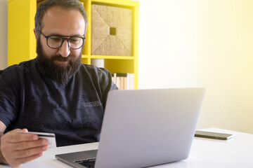 Online shopping, happy, young, bearded man holding credit card online shopping on his laptop. E-commerce, spending money, internet banking.