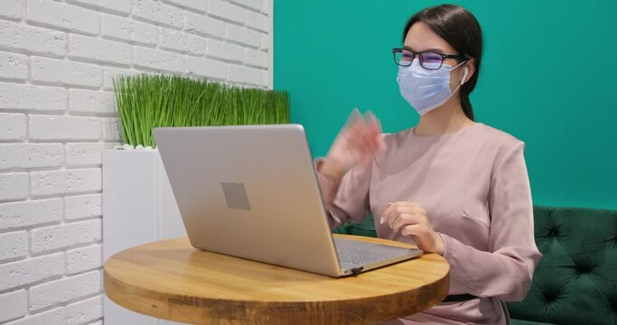 Young woman with face mask works in coworking after lockdown, have video call touches an earphone and talks to colleagues using a laptop gestures with hands