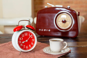 An alarm clock of red color, a white small cup of coffee are in the background of the radio on the...