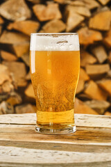 Glass of light beer on a blurred background of firewood. Full glass of fresh golden beer with small...