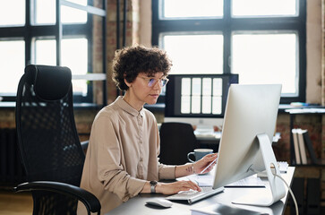 Serious young businesswoman in eyeglasses sitting at office desk in front of computer monitor and typing on computer key
