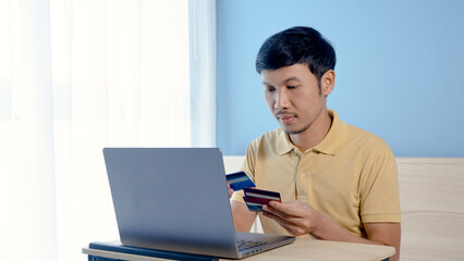 Handsome Asian man, wearing a yellow shirt sitting on a white bed, has a serious look on his face when he chooses a credit card to pay for an order online, on his laptop computer..