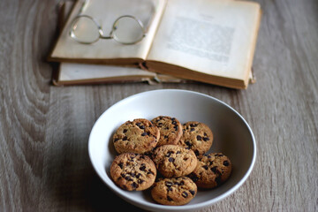 Bowl of strawberries and blueberries, open books with reading glasses, plate of chocolate chip cookies and basket of apples one the table. Selective focus.