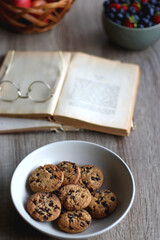 Bowl of strawberries and blueberries, open books with reading glasses, plate of chocolate chip cookies and basket of apples one the table. Selective focus.