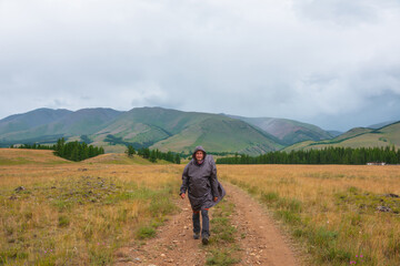 Satisfied tourist returns from mountains in overcast. Happy man in raincoat walks through hills and forest in bad weather. Traveler goes towards adventure. Hiker and mountain range under cloudy sky.