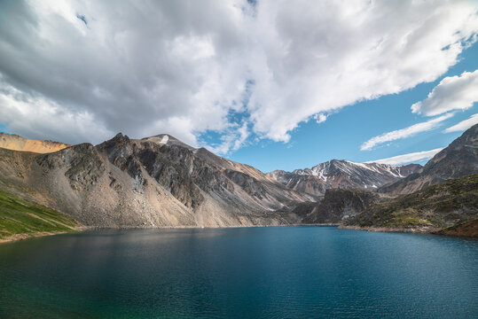 Deep mountain lake of phantom blue color among high mountains with pointed peak in changeable weather. Wonderful dramatic view to deep blue mountain lake among sunlit sharp rocks under cloudy sky. © Daniil