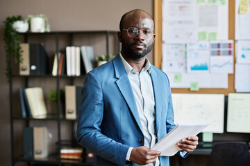 Portrait of African confident businessman in eyeglasses and blue suit looking at camera while working with documents at office