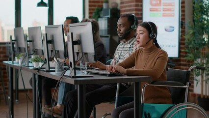 Asian woman with using headset to help clients at call center, talking to people about telemarketing. Employee in wheelchair working at customer assistance service. Tripod shot. chronic health