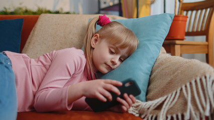 Fototapeta premium Child toddler hold smartphone watching funny cartoons, chatting with friends lying on couch alone at home. Little one school kid girl using mobile phone technology device. Children tech addiction
