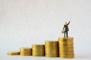Miniature people: businessman  figure raise your hand and holding a bag standing on stack of gold...