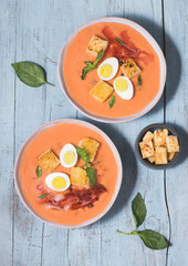 Spanish salmorejo soup, tradition Andalusian
cold tomato soup topped with serrano ham (jamon), boiled egg, bread and  pepper on the blue wooden background. Summer recipes, menu