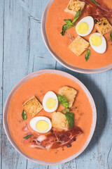 Spanish salmorejo soup, tradition Andalusian
cold tomato soup topped with serrano ham (jamon), boiled egg, bread and  pepper on the blue wooden background. Summer recipes, menu, close-up