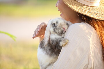 Adorable young rabbit and woman sit together outdoor. Owner care little cute bunny on her laps in forest. Woman wear long sleeves light brown dress sitting and holds flower of grass in relax and calm.