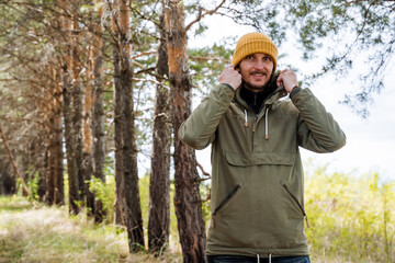 The guy is smiling standing in the forest, a hat on his head, a warm windbreaker from the rain, a man puts a hood on his head, a tourist on a hike in a pine forest.