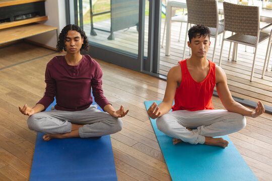 Diverse gay male couple practicing yoga and meditating together in the living room at home