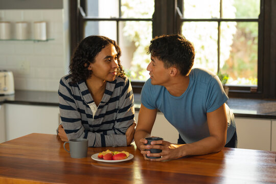Diverse gay male couple holding coffee cups looking at each other in the kitchen at home