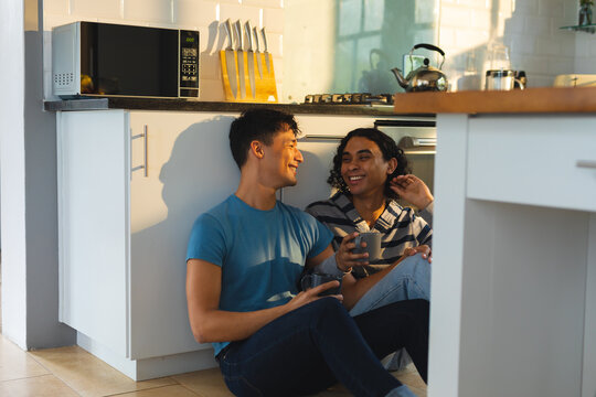 Diverse gay male couple smiling looking at each other sitting on the floor in the kitchen at home
