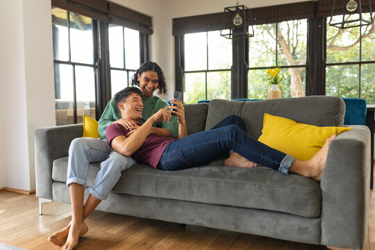 Diverse gay male couple smiling while using smartphone sitting together on the couch at home