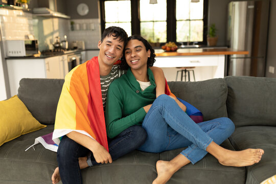 Diverse gay male couple with lgbt flag wrapped round their back sitting together on the couch