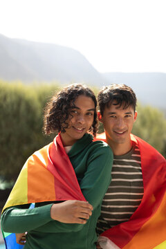 Portrait of diverse gay male couple with lgbt flag wrapped round their back smiling together