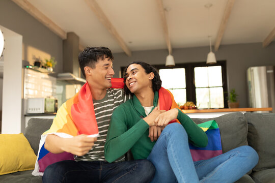 Diverse gay male couple with lgbt flag wrapped round their back sitting together on the couch