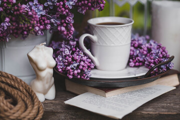 A beautiful postcard. A white coffee cup with a saucer, candles and a vase with a bouquet of purple lilac. The color 2022 is very peri. Beautiful still life. Spring time. The concept of 