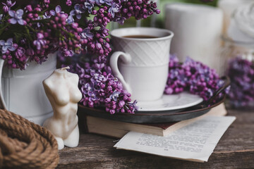 A beautiful postcard. A white coffee cup with a saucer, candles and a vase with a bouquet of purple lilac. The color 2022 is very peri. Beautiful still life. Spring time. The concept of 