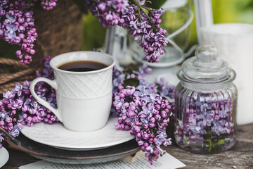 Obraz na płótnie Canvas A beautiful postcard. A white coffee cup with a saucer, candles and a vase with a bouquet of purple lilac. The color 2022 is very peri. Beautiful still life. Spring time. The concept of 