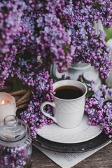 Obraz na płótnie Canvas A beautiful postcard. A white coffee cup with a saucer, candles and a vase with a bouquet of purple lilac. The color 2022 is very peri. Beautiful still life. Spring time. The concept of 