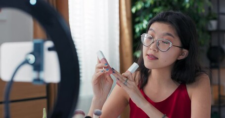 Female beauty blogger filming daily makeup routine tutorial using ring light with phone on tripod. Influencer with dark skin live streaming about cosmetics, comparing two shades of matte lipsticks.