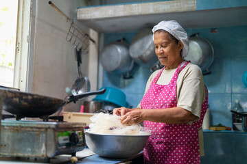 Portrait of older Asian woman wearing chef hat and pink apron cooking Thai food in kitchen at home....